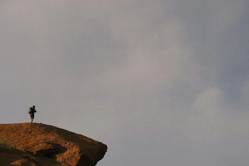 A lone hiker stands on the edge of a rocky cliff under an expansive gray sky