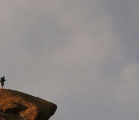A lone hiker stands on the edge of a rocky cliff under an expansive gray sky