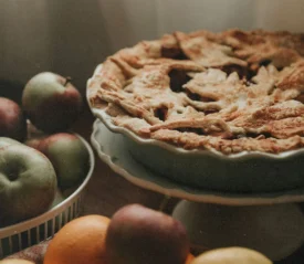 A freshly baked pie with a golden, textured crust sits on a stand next to a bowl of apples and assorted fruit
