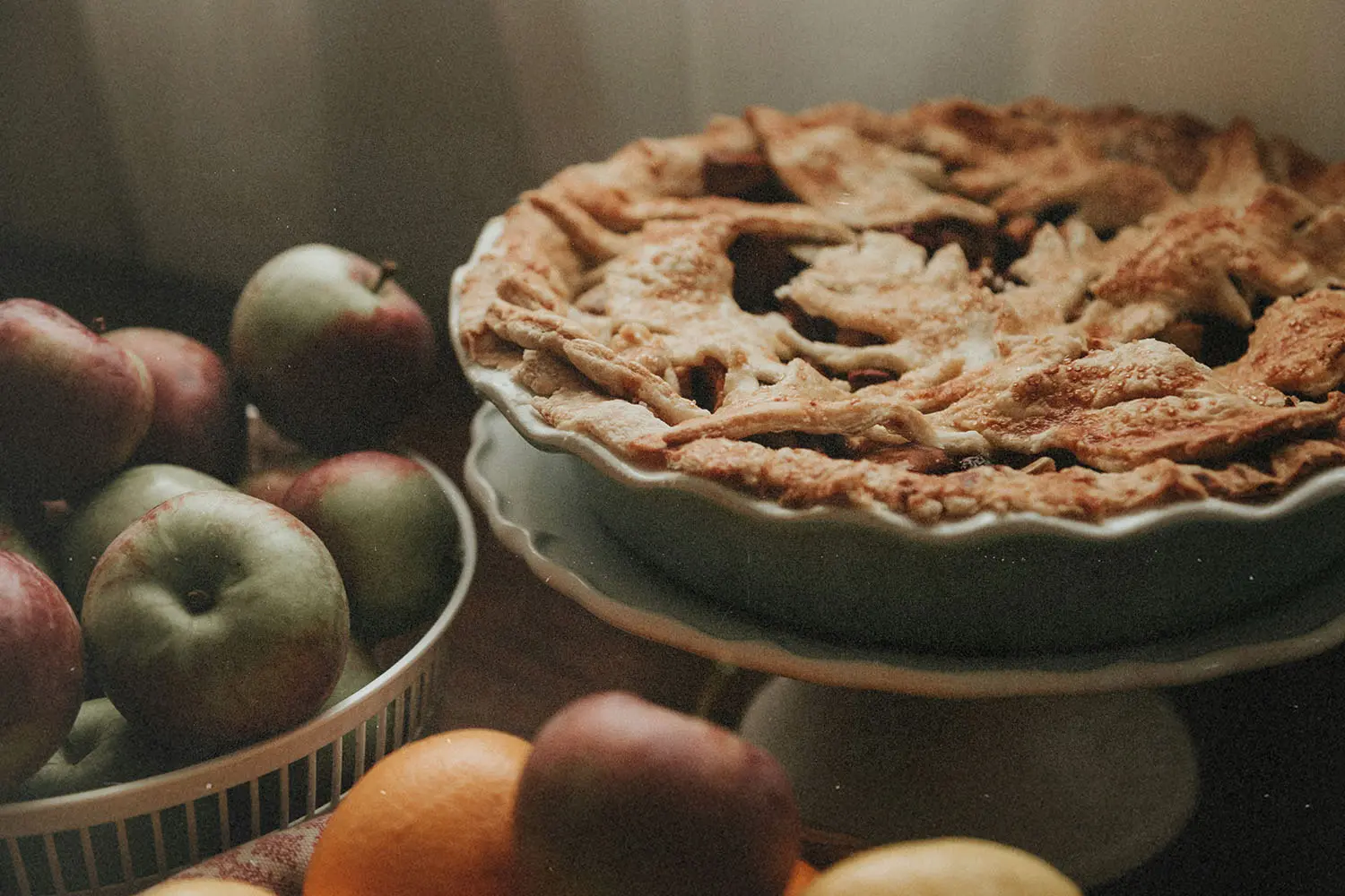 A freshly baked pie with a golden, textured crust sits on a stand next to a bowl of apples and assorted fruit
