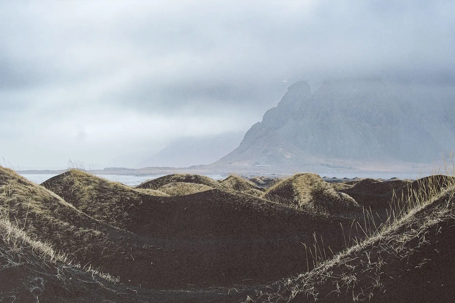 Wind-shaped black sand dunes under a cloudy sky with a mist-covered mountain in the distance