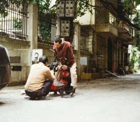Two people working together to repair a red motorbike on a narrow residential street lined with cars, plants, and aging buildings