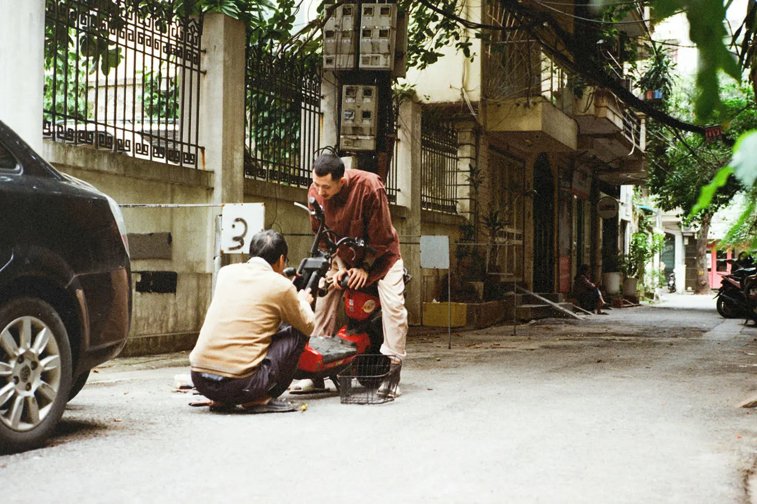 Two people working together to repair a red motorbike on a narrow residential street lined with cars, plants, and aging buildings