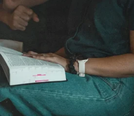 A person sits with an open book resting on their lap, their hand placed on the pages, wearing a watch and denim jeans