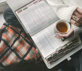 A person sits by a window with a warm drink, reading an open book filled with colorful tabs and highlighted text, with a patterned blanket nearby