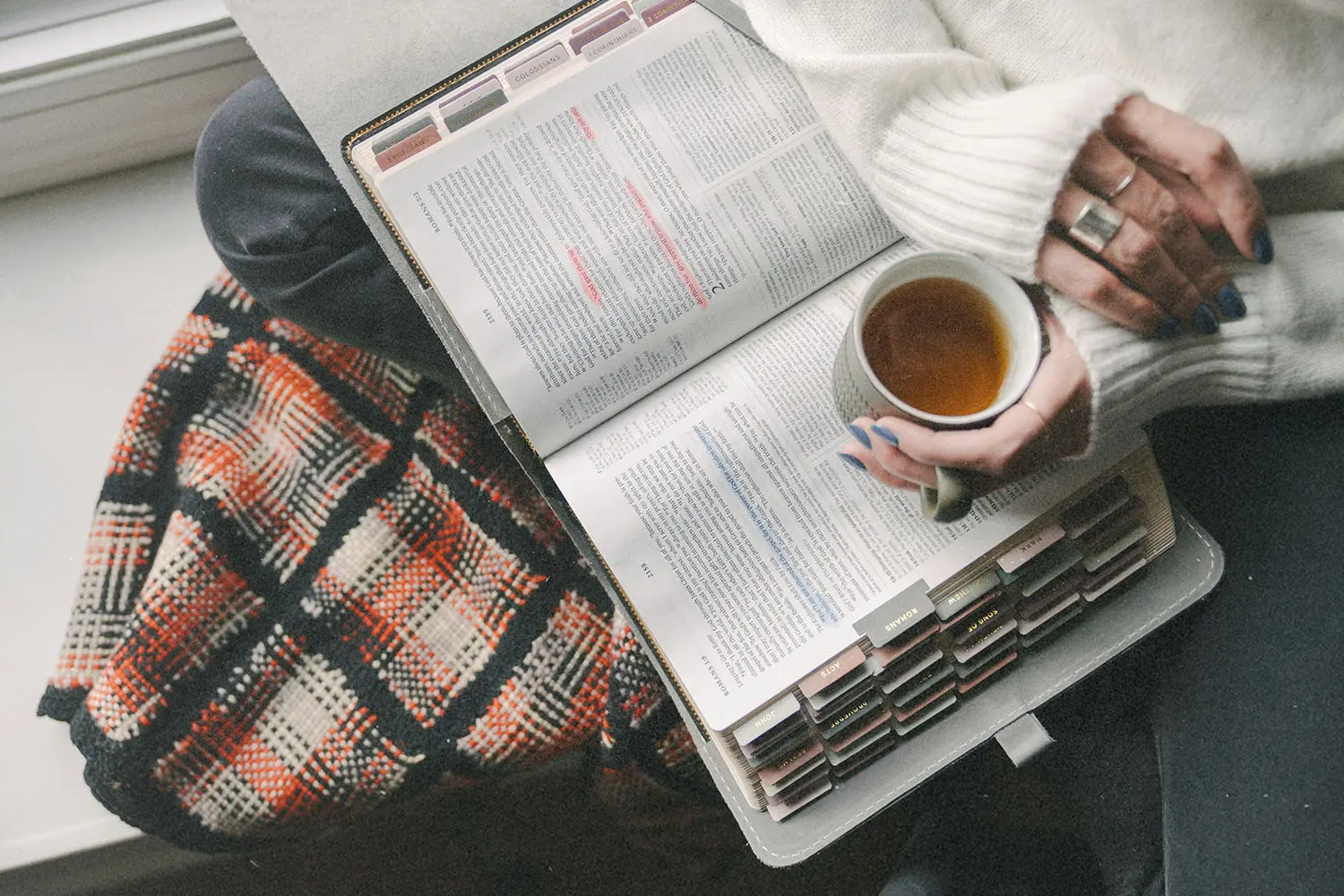 A person sits by a window with a warm drink, reading an open book filled with colorful tabs and highlighted text, with a patterned blanket nearby