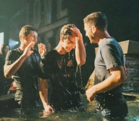 A group of people stand in a shallow pool during a baptism ceremony, water dripping as one person touches their forehead