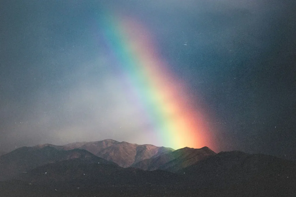 A bright rainbow arcs downward toward a mountain range under a dark, cloudy sky