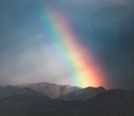 A bright rainbow arcs downward toward a mountain range under a dark, cloudy sky