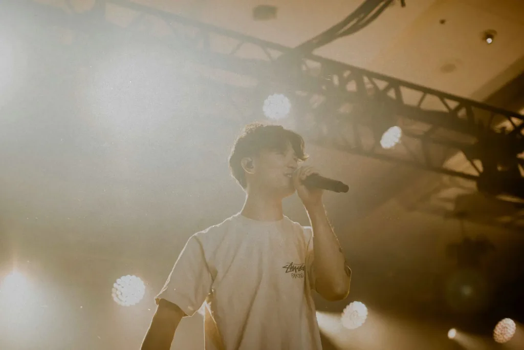 A performer holds a microphone on a brightly lit stage, surrounded by warm spotlights and overhead rigging