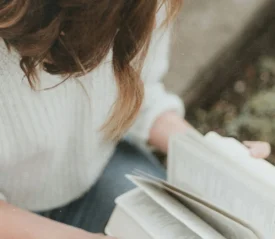 A person in a white sweater reads a book outdoors, holding the pages open across their lap