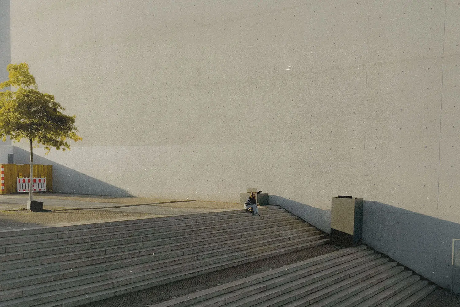 A lone person sits on wide concrete steps beside a large, plain wall, with a small tree casting a long shadow nearby
