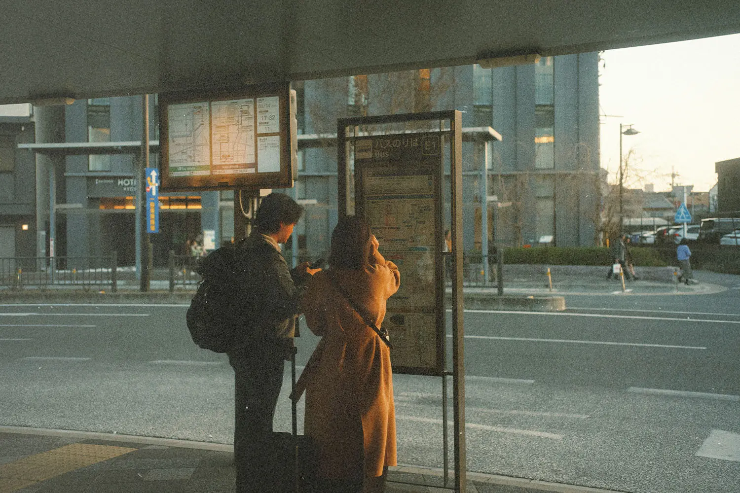 Two people stand at a bus stop examining a posted route map as evening sunlight falls across the street