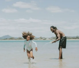 A child runs through shallow water at the beach as an adult playfully splashes water toward them under a bright sky