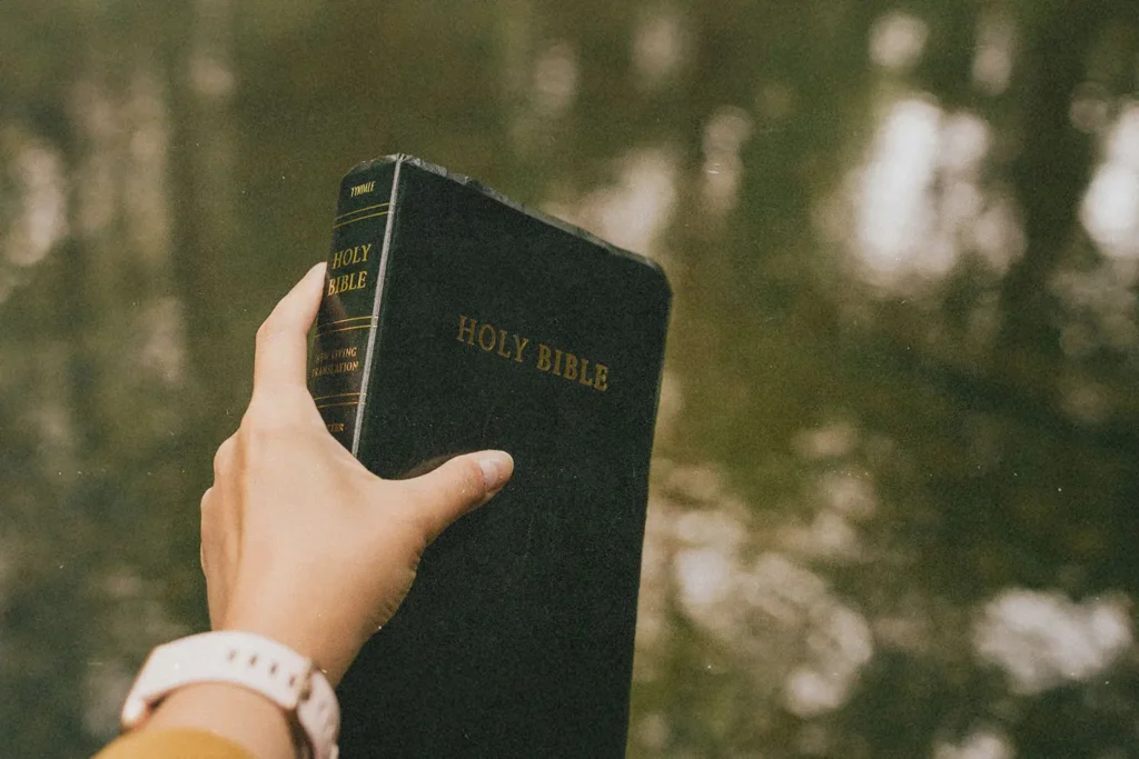 A hand holds up a black Holy Bible outdoors, with water reflecting soft natural light in the background
