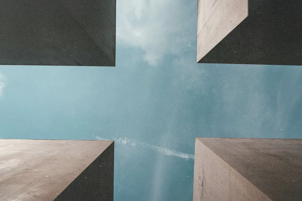 Upward view between four tall concrete structures forming a cross-shaped opening to the sky