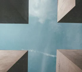 Upward view between four tall concrete structures forming a cross-shaped opening to the sky