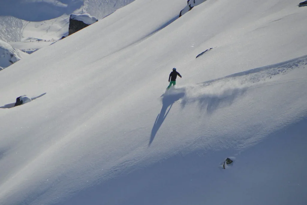 A skier glides down a wide, snow-covered mountain slope, leaving a trail of powder behind under bright sunlight