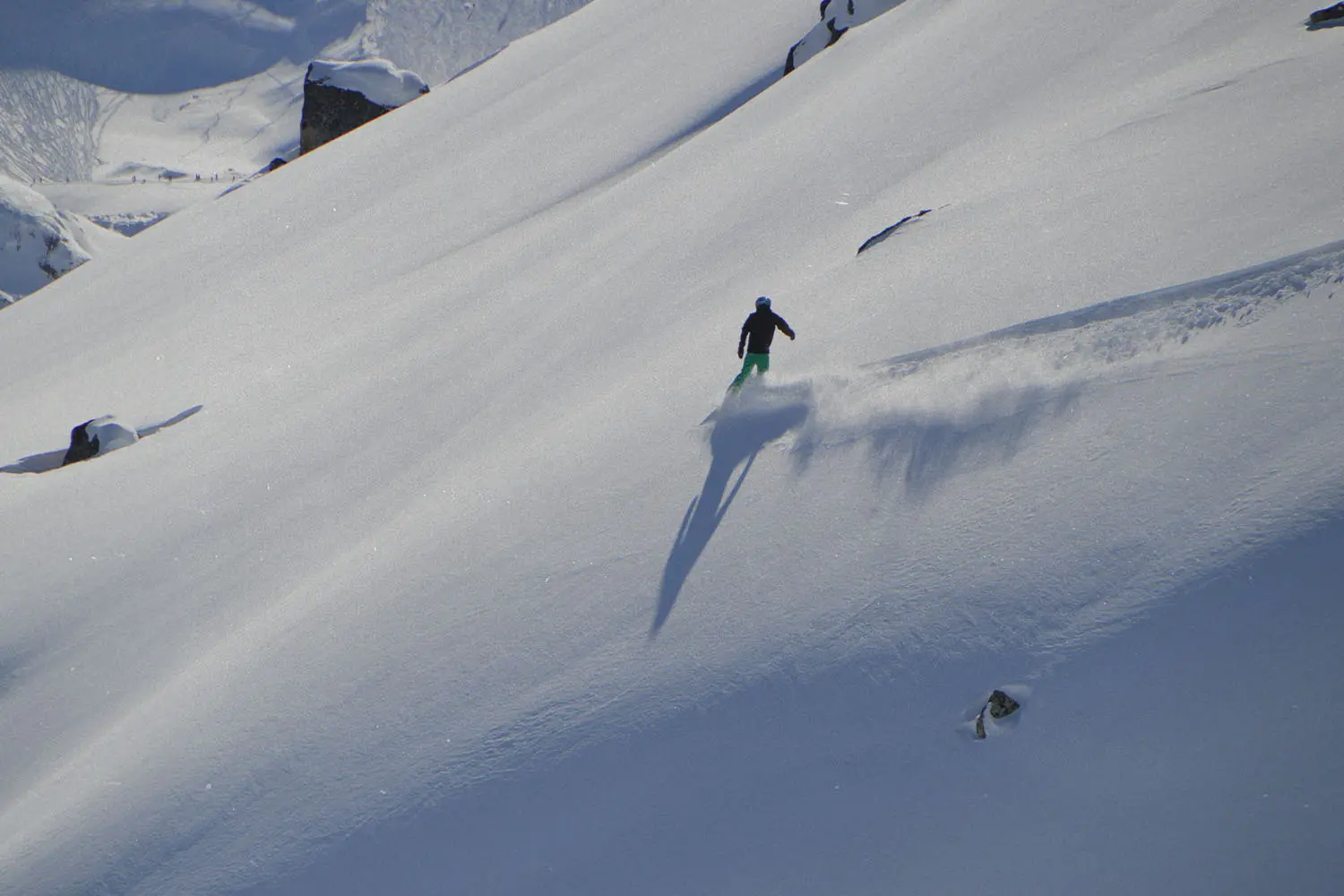 A skier glides down a wide, snow-covered mountain slope, leaving a trail of powder behind under bright sunlight
