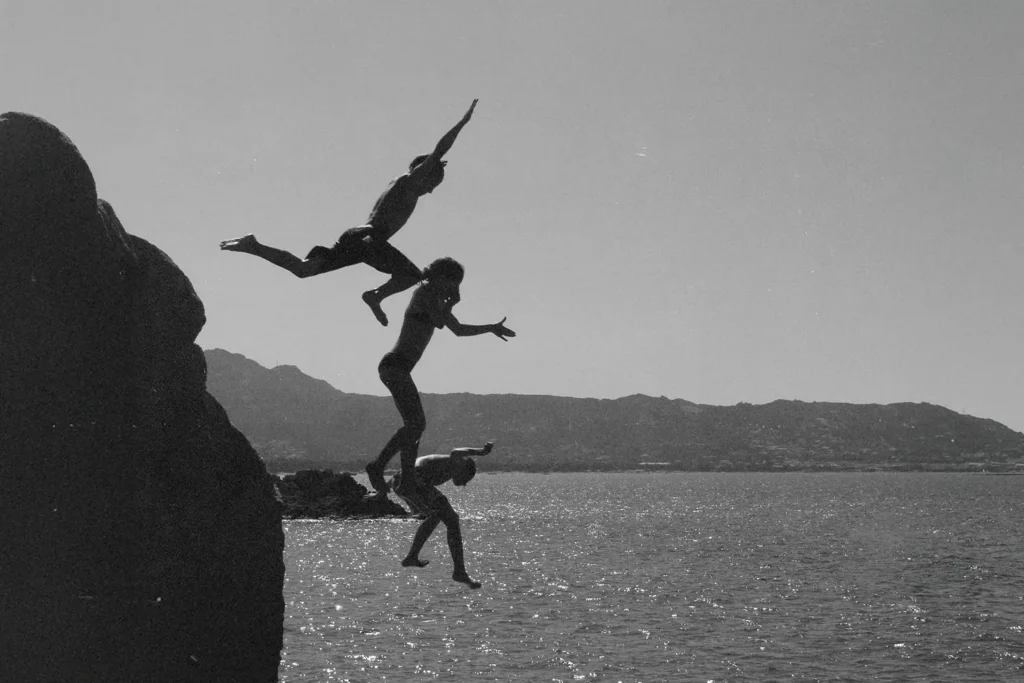 Three people leap from a rocky cliff into the water below, captured mid‑air in a black‑and‑white scene