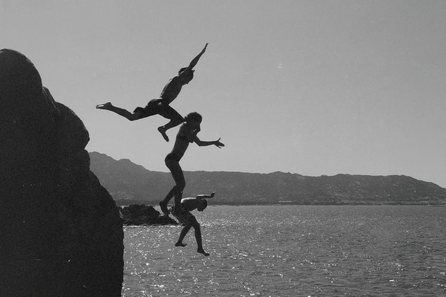 Three people leap from a rocky cliff into the water below, captured mid‑air in a black‑and‑white scene