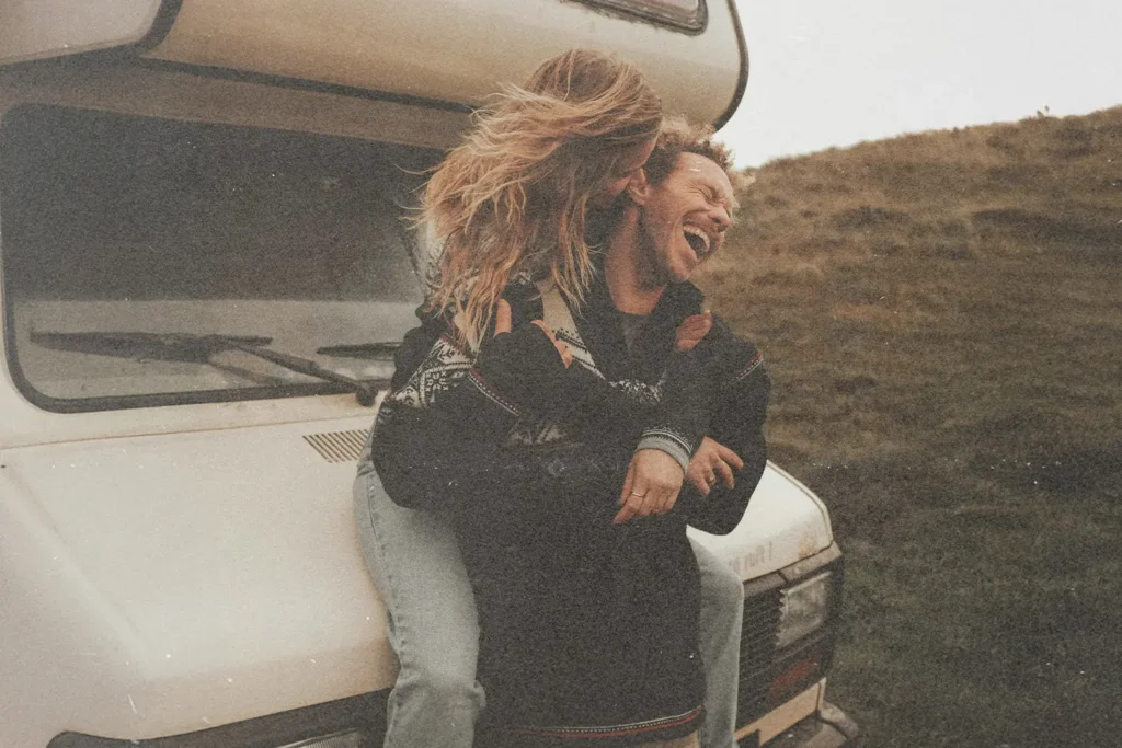 Two people sitting on the front of a camper parked on grassy terrain, with rolling hills in the background under an overcast sky.
