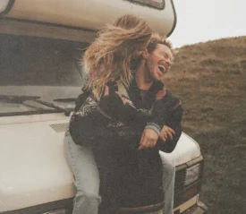 Two people sitting on the front of a camper parked on grassy terrain, with rolling hills in the background under an overcast sky.