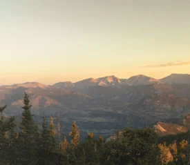 A mountain range glows in warm sunset light, with trees in the foreground and layered peaks stretching into the distance