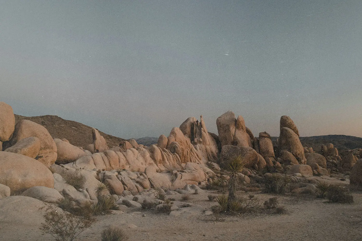 A desert landscape filled with rounded rock formations and sparse shrubs under a pale early‑evening sky