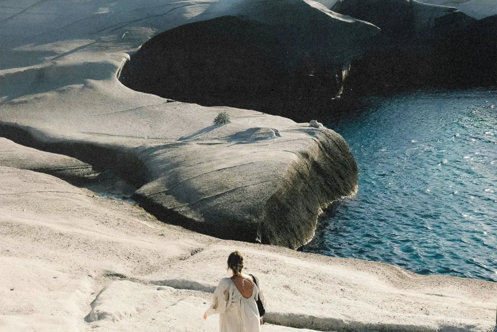 A person walks down pale, sculpted rock formations toward the edge of bright blue water in a sunlit coastal landscape