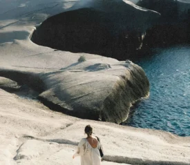 A person walks down pale, sculpted rock formations toward the edge of bright blue water in a sunlit coastal landscape