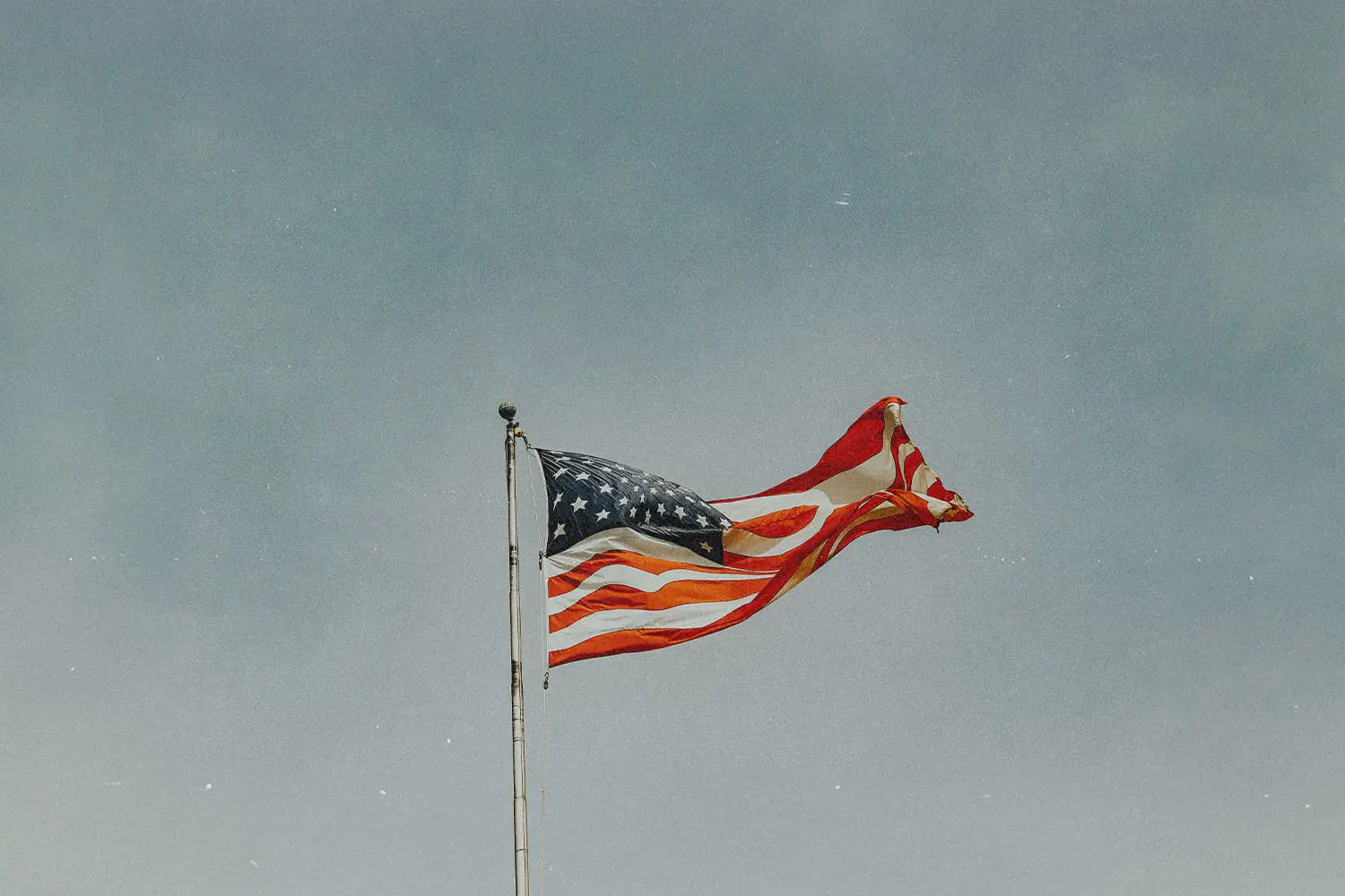 Weathered U.S. flag rippling on a tall pole beneath a cloudy sky