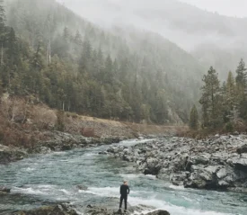 Man standing at a river bank looking out to a forest