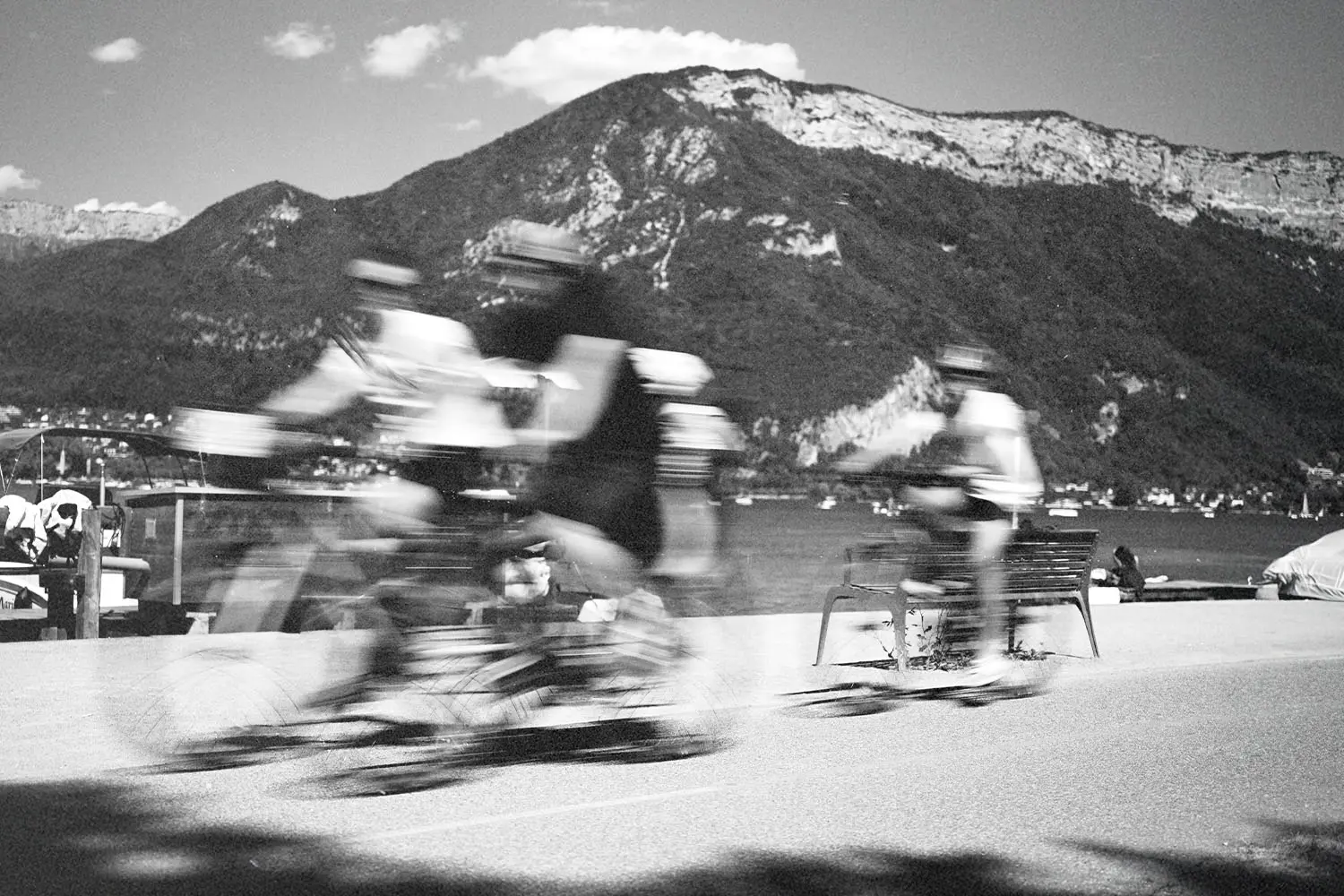 Motion-blurred cyclists moving along a waterfront trail with towering mountains behind them