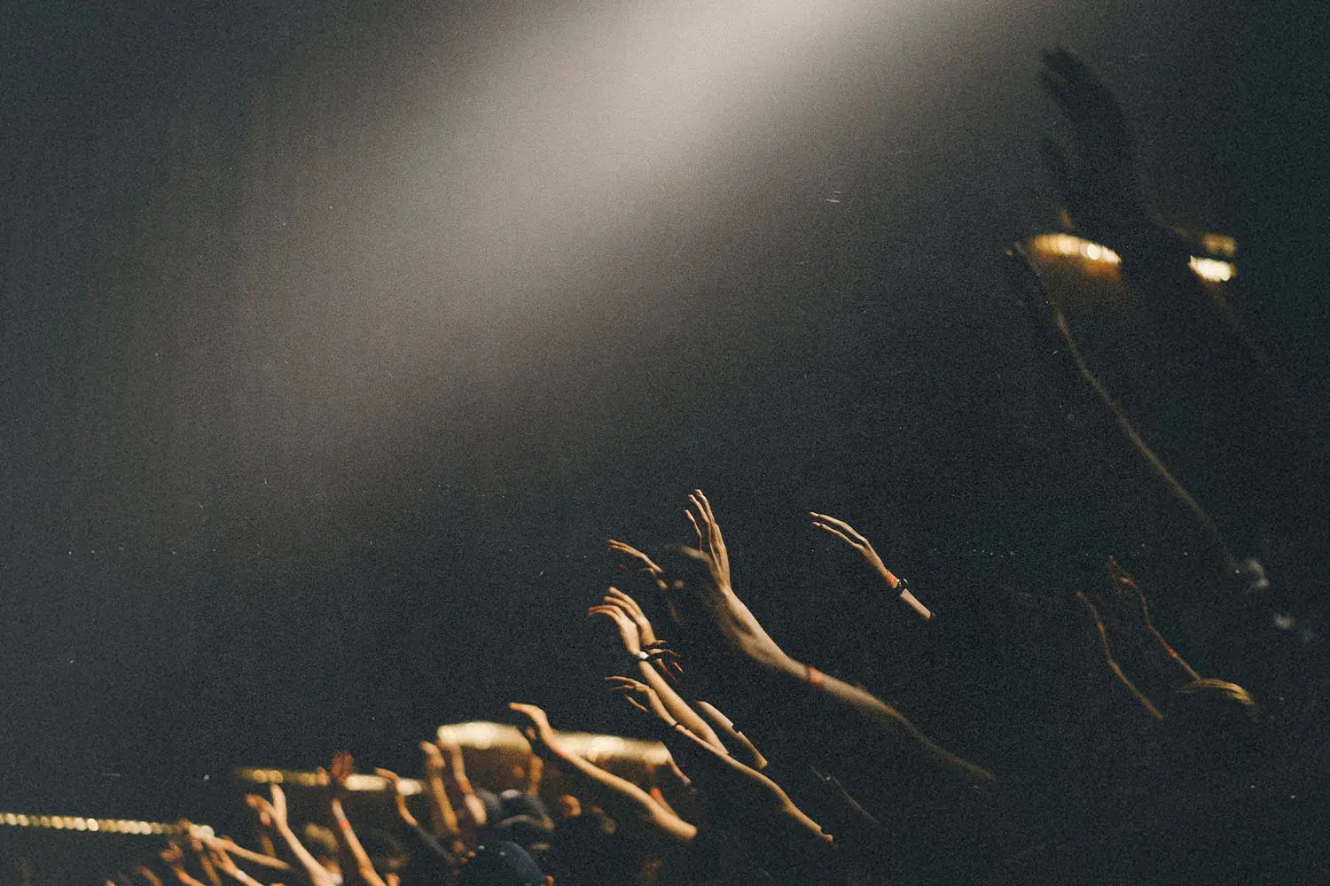 Numerous raised hands reach upward toward a bright spotlight in a dark room