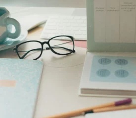 A workspace with glasses, notebooks, a planner, a keyboard, and a ceramic mug arranged neatly on a light-colored desk