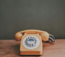 A vintage yellow rotary telephone sits on a wooden surface in front of a muted green wall