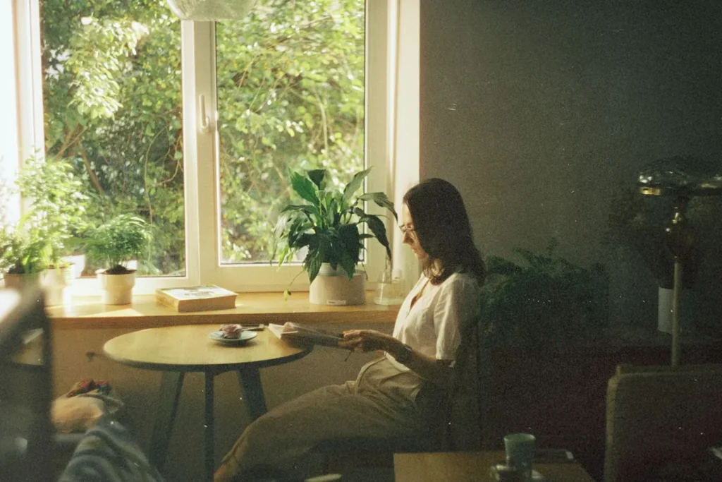 Woman reading at a small table beside a bright window surrounded by potted plants