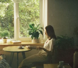 Woman reading at a small table beside a bright window surrounded by potted plants