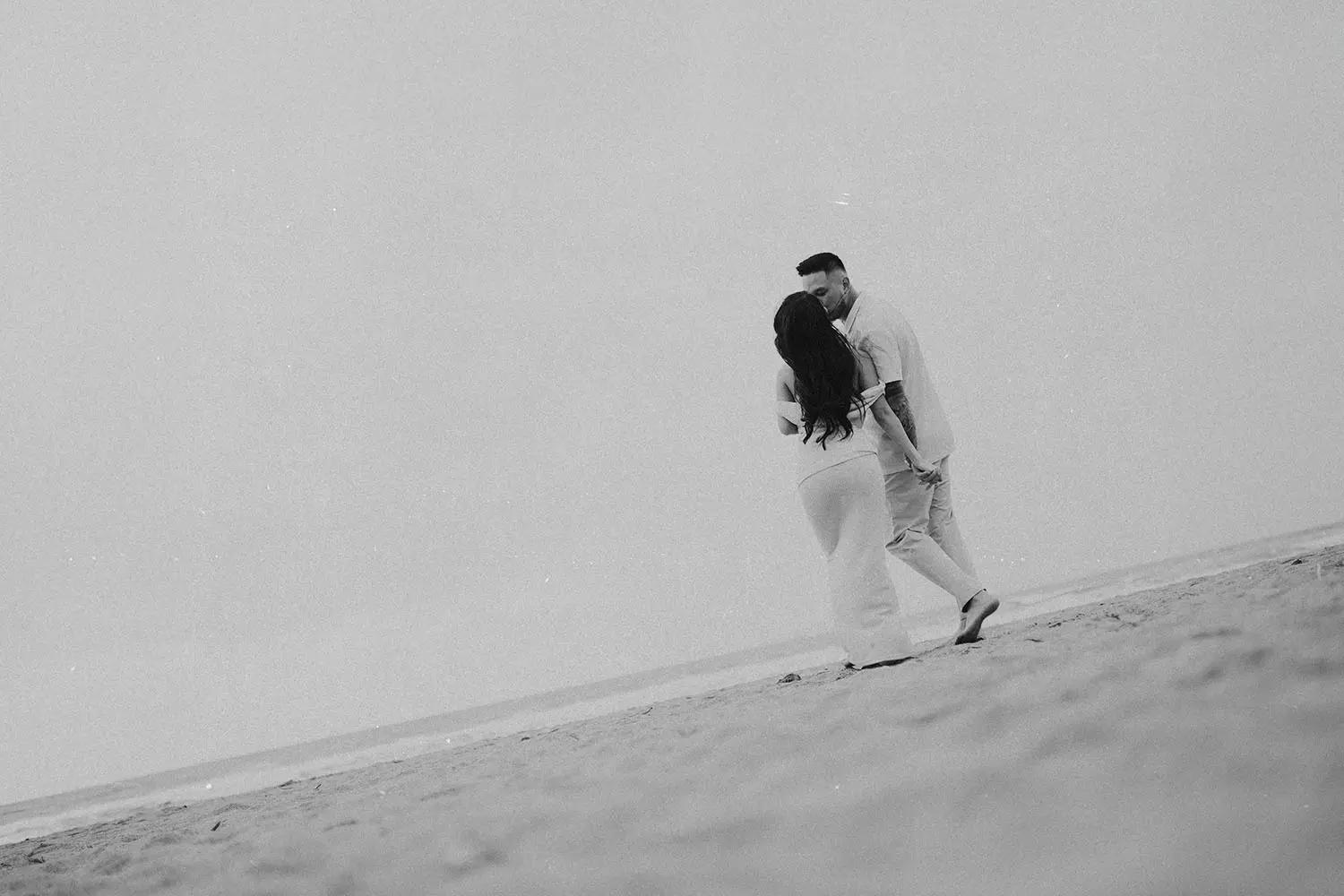 A couple stands close together on a quiet beach, holding hands as they face each other beside the shoreline