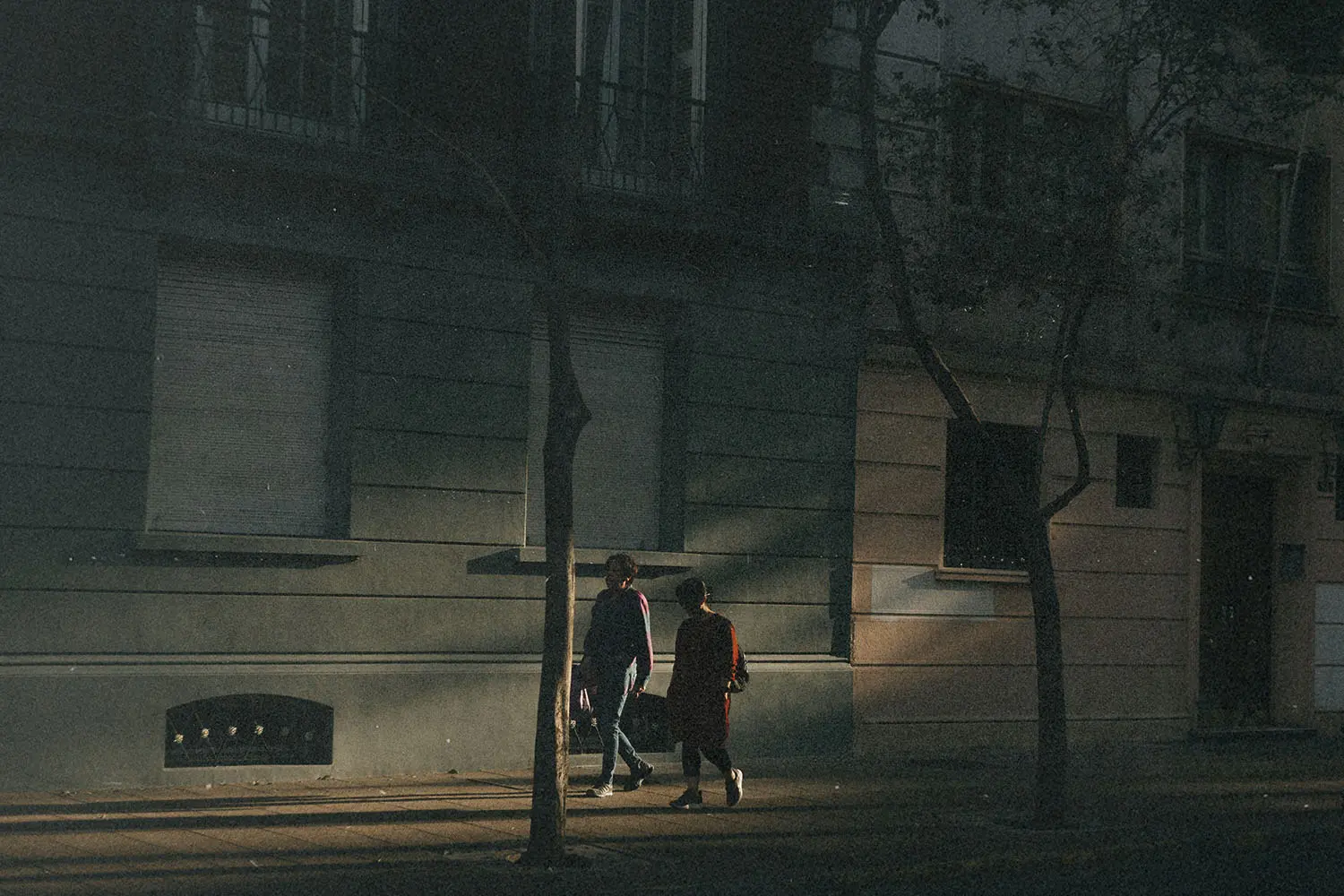Two people walk side by side down a dimly lit urban street, illuminated by a beam of late‑day sunlight falling across the sidewalk