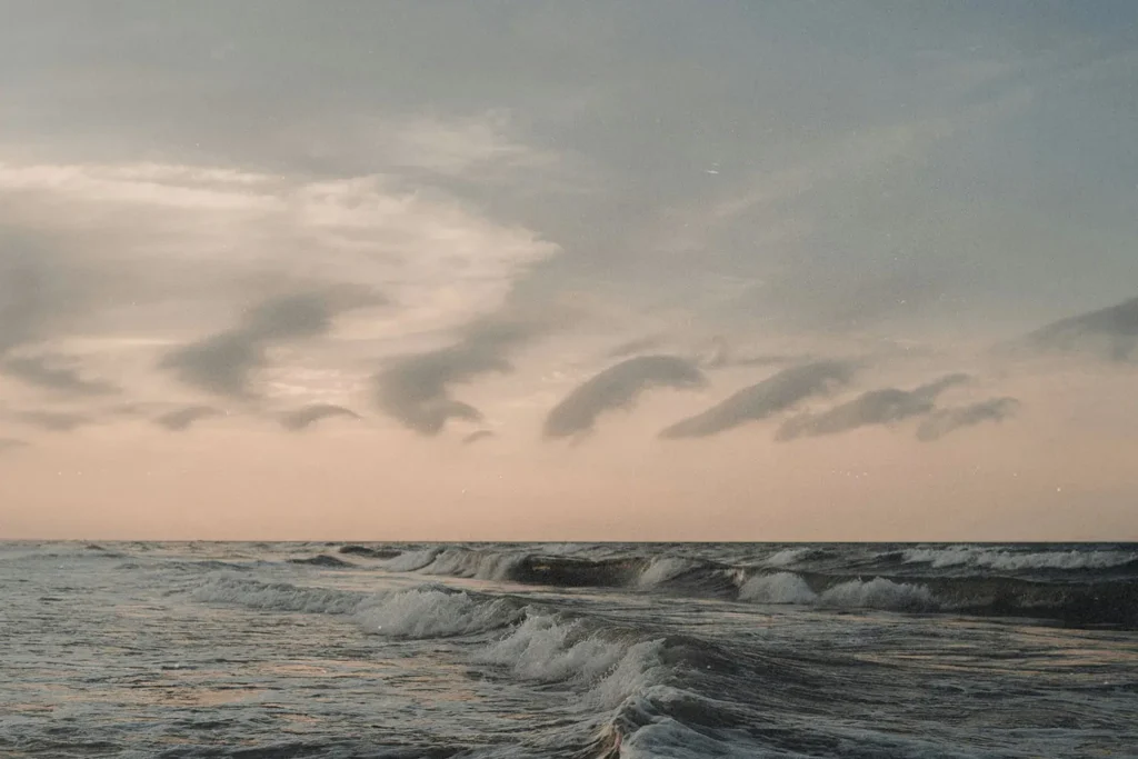 Ocean waves rolling toward the shore beneath a pastel sky with soft, curved clouds at sunrise or sunset