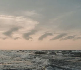 Ocean waves rolling toward the shore beneath a pastel sky with soft, curved clouds at sunrise or sunset