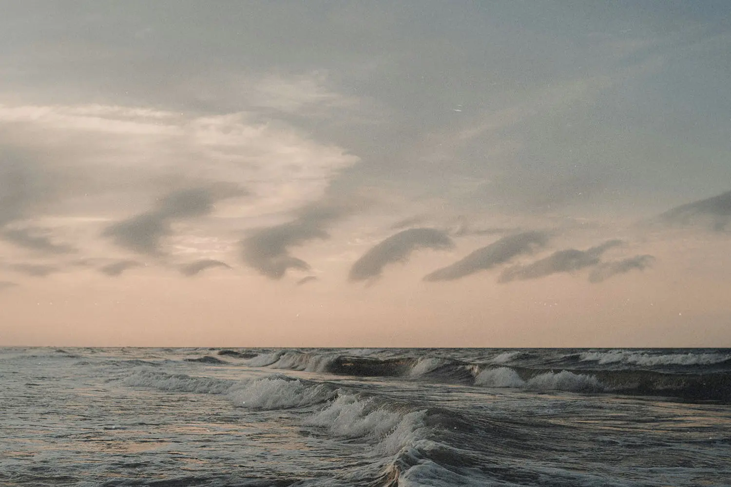 Ocean waves rolling toward the shore beneath a pastel sky with soft, curved clouds at sunrise or sunset