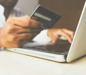 A person holds a credit card in one hand while using the other hand to type on a laptop, suggesting an online payment or purchase