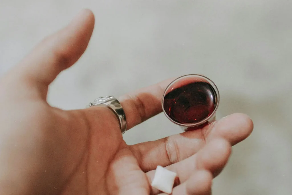 A hand holds a small cup of red communion juice with a white communion wafer resting near the fingertips