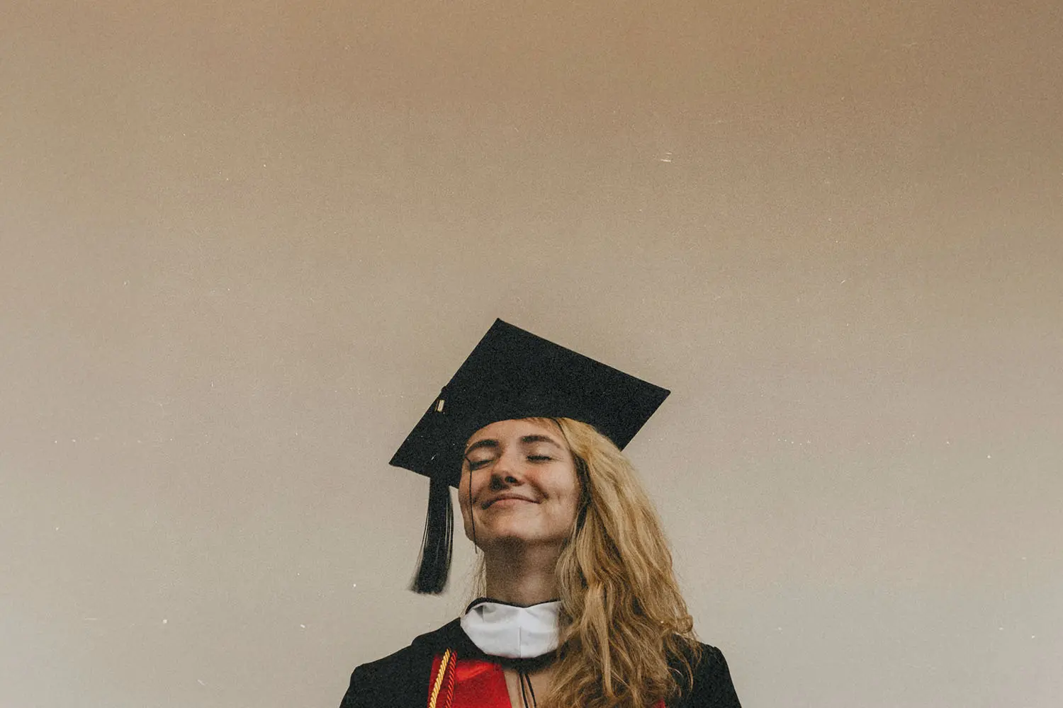 A graduate wearing a cap and gown stands against a plain beige background