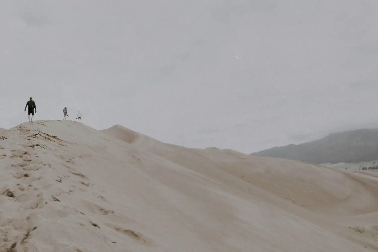 Three people walking up a steep sand dune, leaving footprints in behind