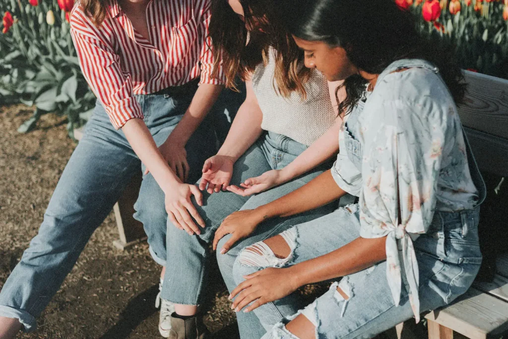 Three people sit together outdoors, hands lightly touching in a supportive gesture, surrounded by sunlight and nearby tulips