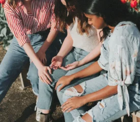 Three people sit together outdoors, hands lightly touching in a supportive gesture, surrounded by sunlight and nearby tulips